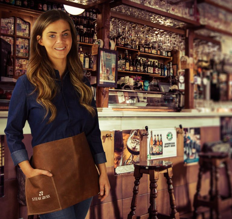 short-waist-bar-apron-with-steal-on-six-embroidery Short Bar Waist Apron On Model In Bar with Steak On Six Logo embroidery